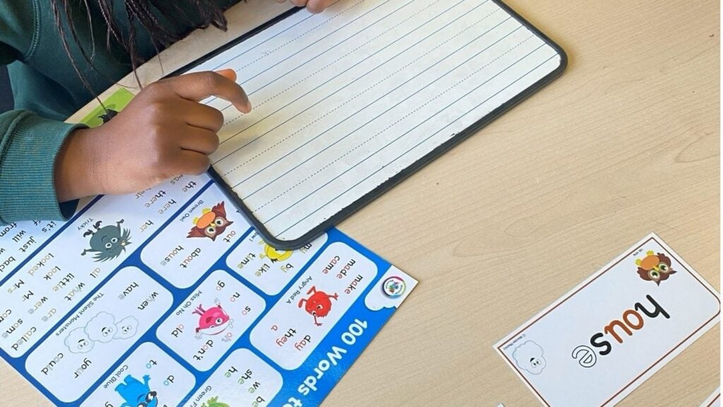 A child writing tricky words on a whiteboard next to the Monster Phonics 100 Words to Learn mat