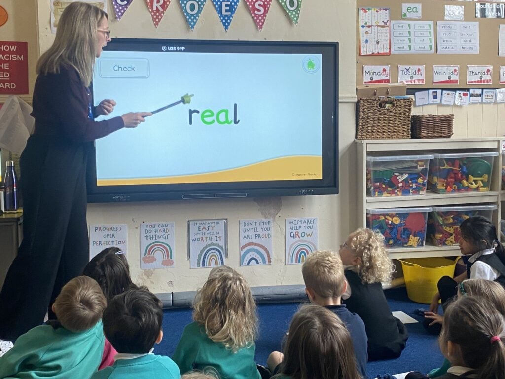 A teacher in a Welsh primary school leading a whole-class phonics lesson, with the word real on the screen